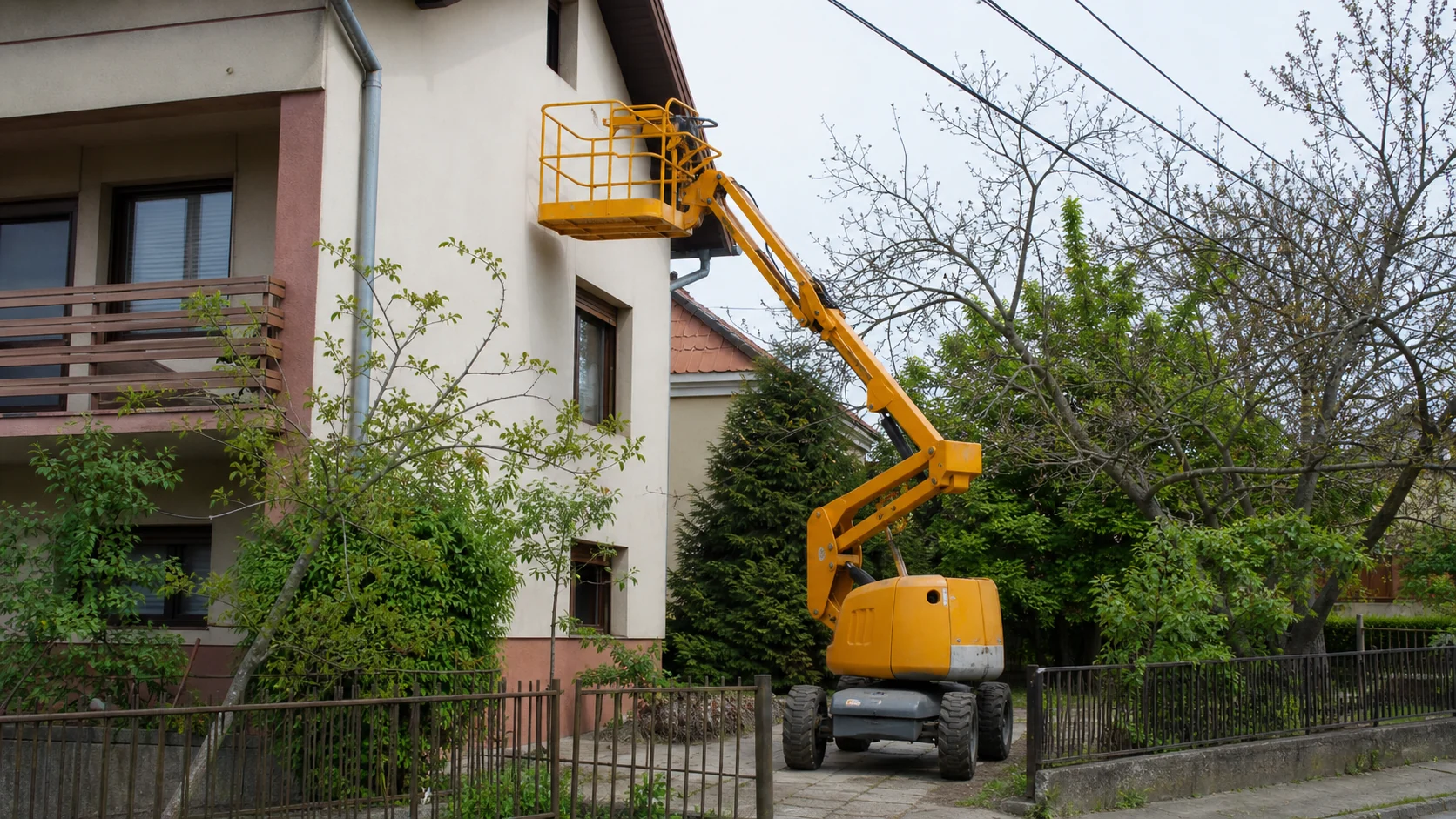 Podnośnik koszowy przy budynku z widocznymi drzewami i przewodami